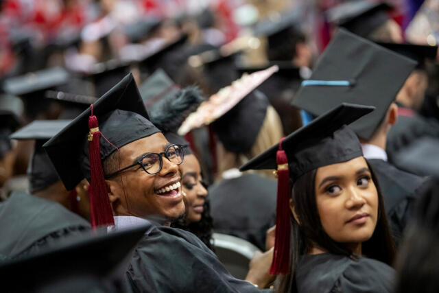 Two graduates facing the camera and smiling over their shoulders in a large group of other graduates facing away from the camera.